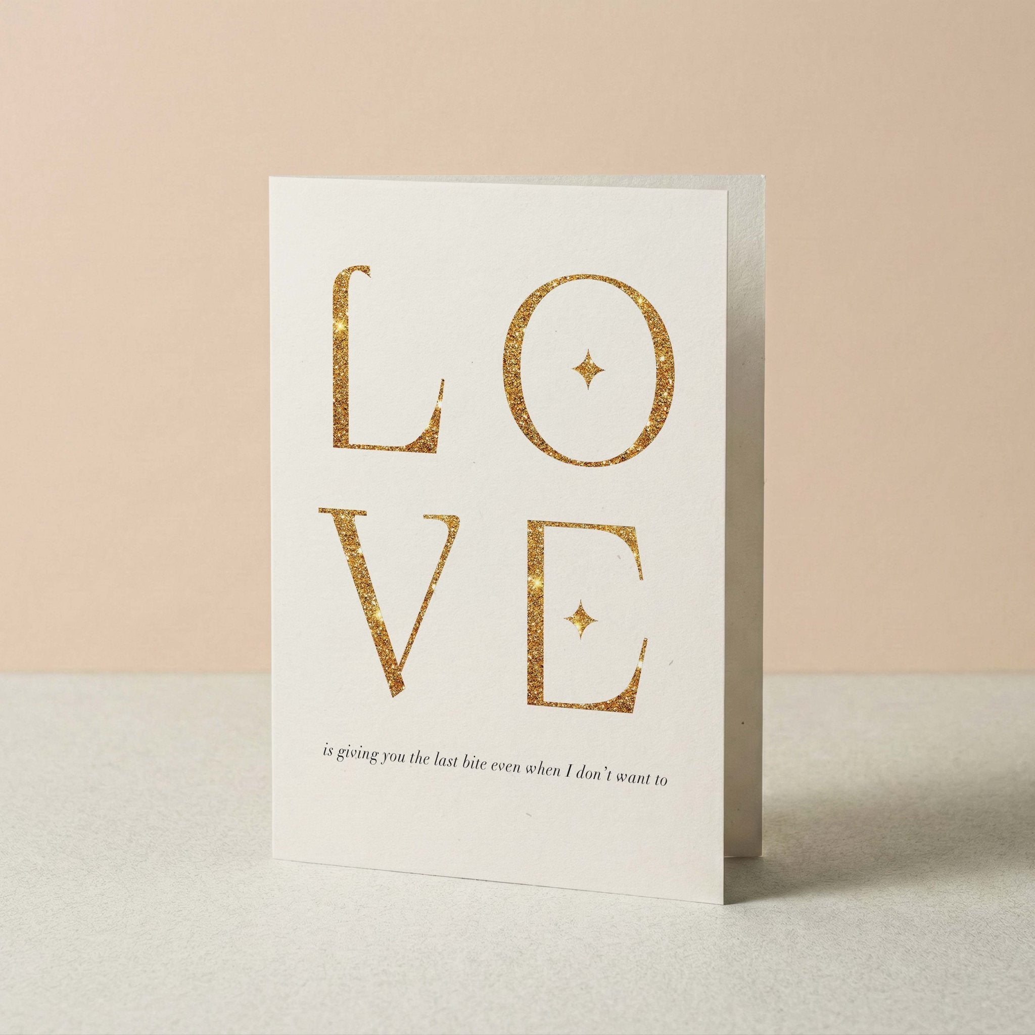 A white greeting card standing upright on a light surface against a soft beige background. The card features the word 'LOVE' in large glittery gold letters, with the 'is giving you the last bite even when I don't want to' in smaller black text underneath.