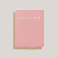 A pink greeting card with the words 'LOVE YOU MORE.' printed in white serif text, shown with a brown kraft envelope on a light gray background.