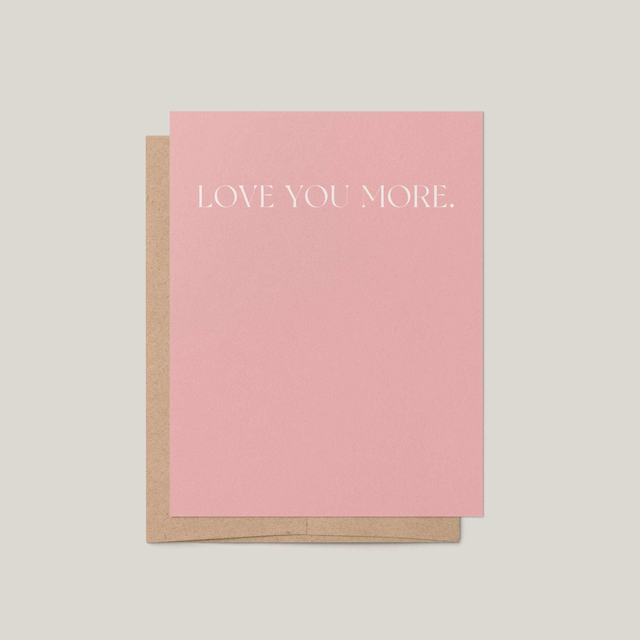 A pink greeting card with the words 'LOVE YOU MORE.' printed in white serif text, shown with a brown kraft envelope on a light gray background.