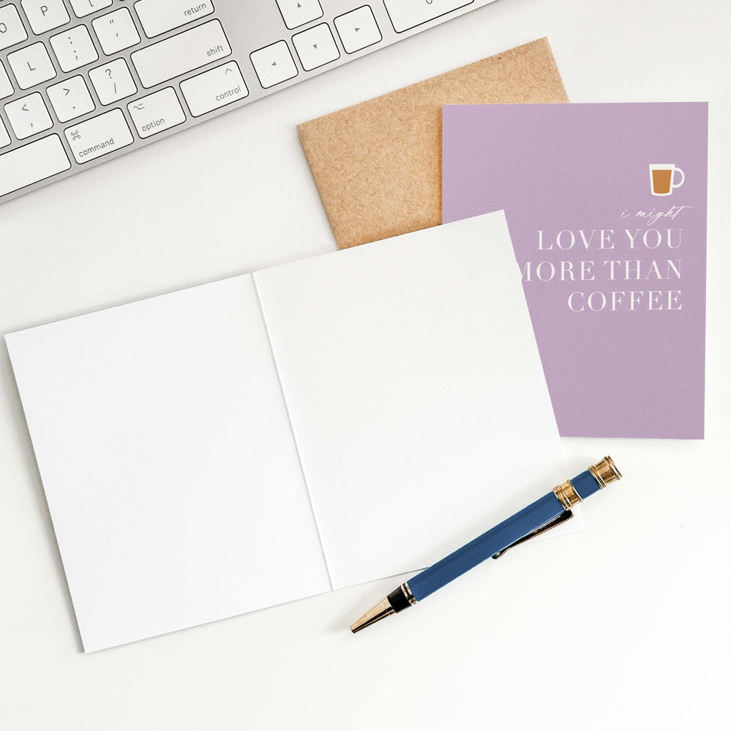Love You More Than Coffee card by Blú Rose shown open with blank interior, kraft envelope and pen on a white desk