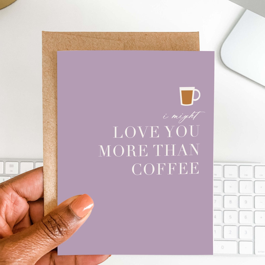Love You More Than Coffee card by Blú Rose held in hand over a white desk with keyboard and kraft envelope