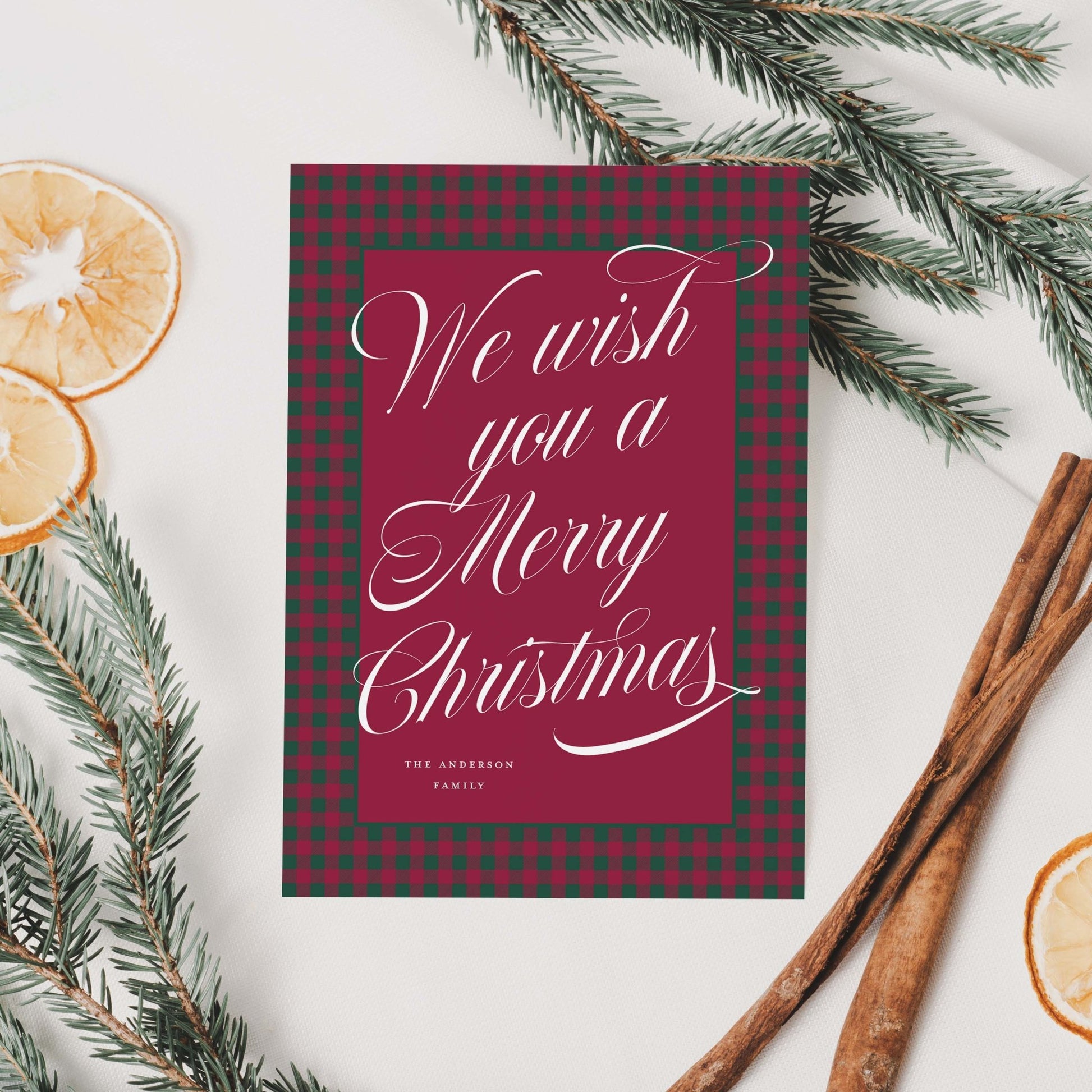 Plaid holiday card surrounded by dried orange slices, pine branches, and ribbon on a neutral background.