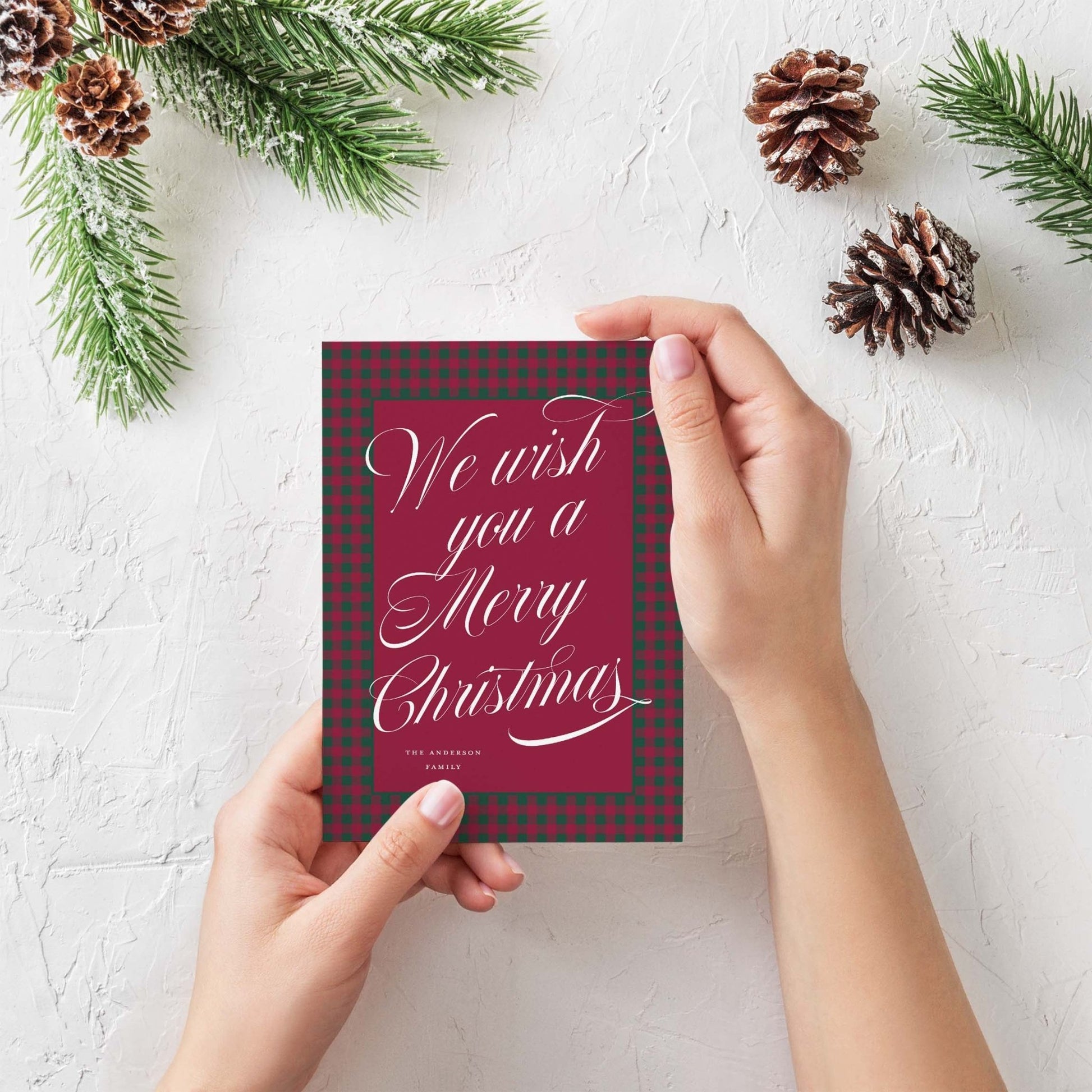 Hand holding the red and green plaid custom holiday card surrounded by pinecones and greenery.