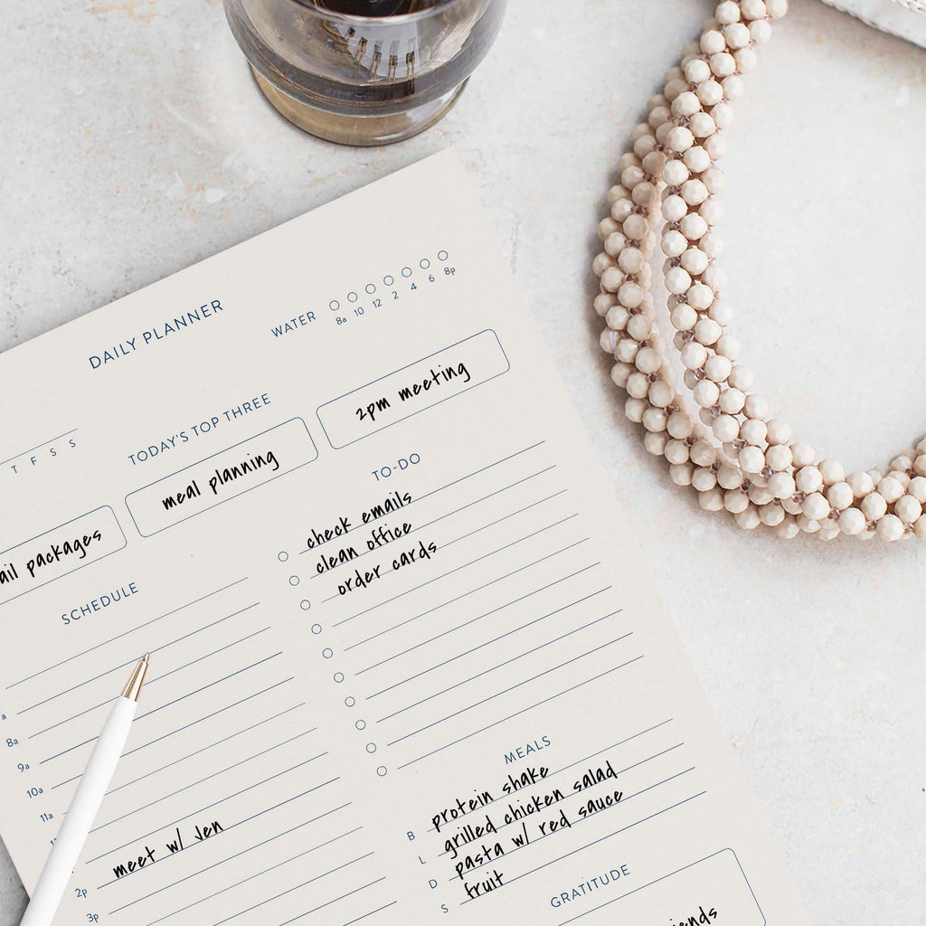 Minimalist Daily Planner notepad by Blú Rose, planner page filled in with handwritten to-dos and meals, styled with a white pen and beaded necklace