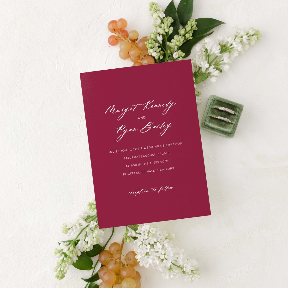 Minimalist Margot wedding invitation by Blú Rose, burgundy card with white script names, styled with white lilacs, grapes, and wedding rings
