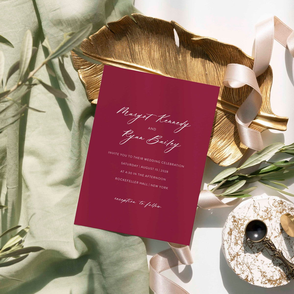 Minimalist Margot wedding invitation by Blú Rose, burgundy card with white script names, styled on a gold leaf dish with sage linen and ribbon