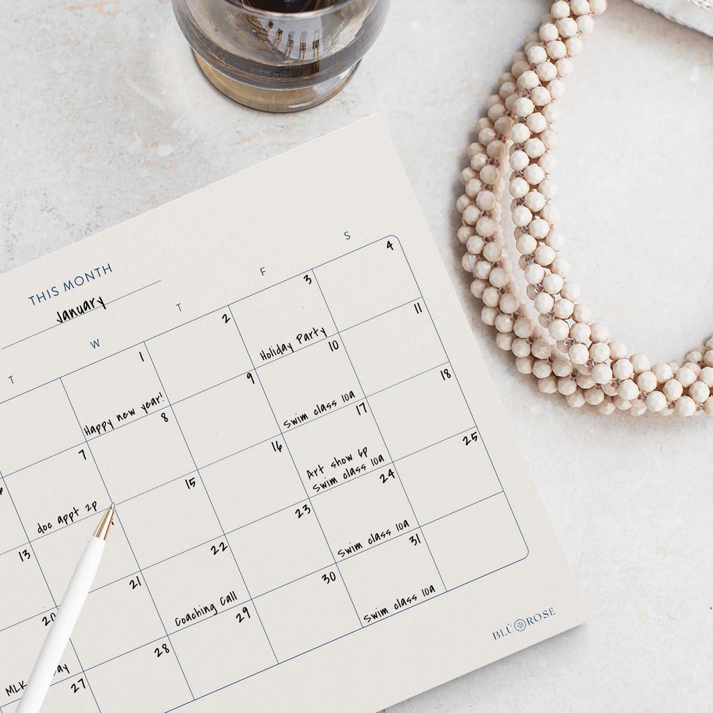 Minimalist Monthly Planner notepad by Blú Rose, monthly calendar filled in with handwritten events, styled with a white pen and beaded necklace