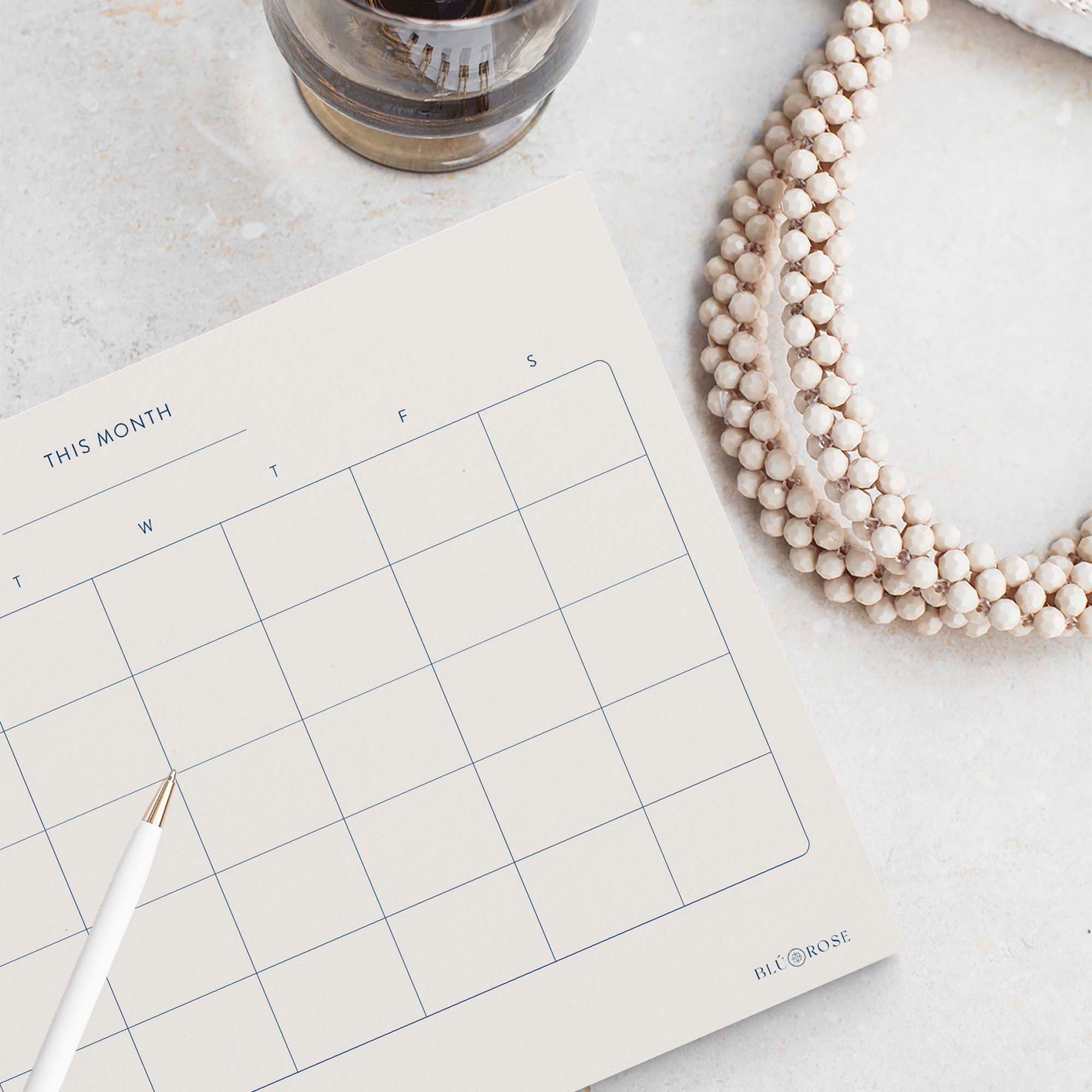 Minimalist Monthly Planner notepad by Blú Rose, blank monthly calendar grid styled with a white pen and beaded necklace
