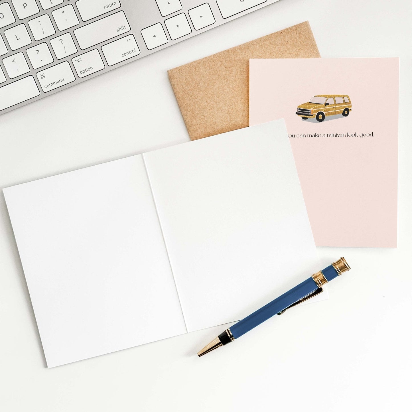 Flat lay of a blush pink greeting card and kraft envelope beside a navy pen on a white surface, styled with minimal stationery accessories.