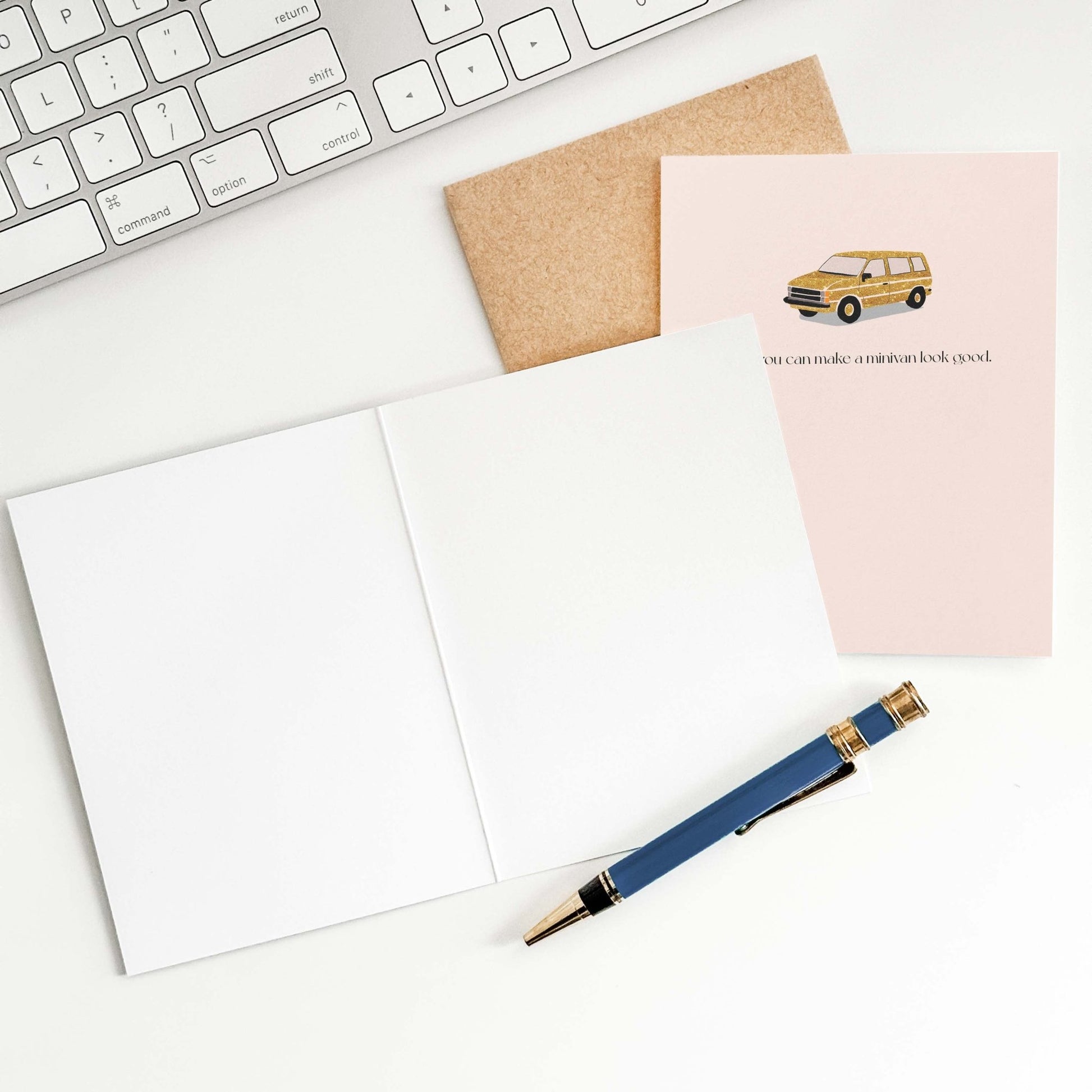 Flat lay of a blush pink greeting card and kraft envelope beside a navy pen on a white surface, styled with minimal stationery accessories.