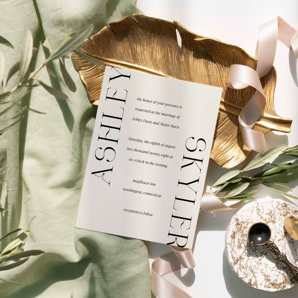 Modern Ashley wedding invitation by Blú Rose, cream card with oversized vertical names, styled on a gold leaf dish with sage linen and ribbon