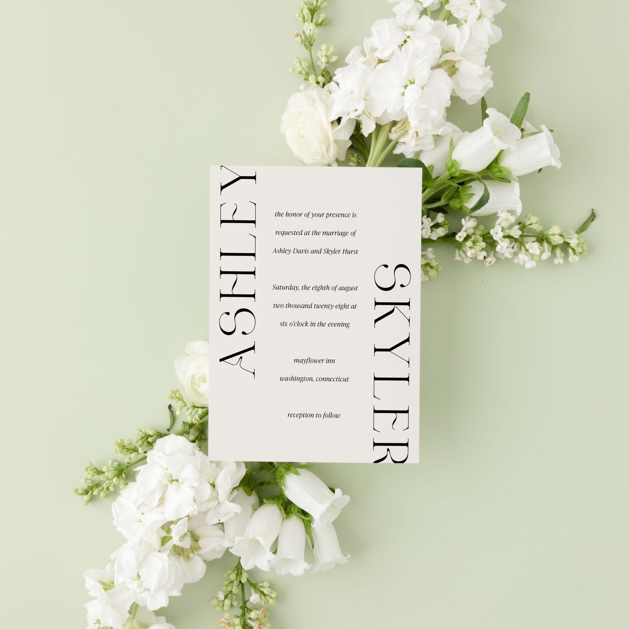 Modern Ashley wedding invitation by Blú Rose, cream card with oversized vertical names, styled on a sage background with white blooms