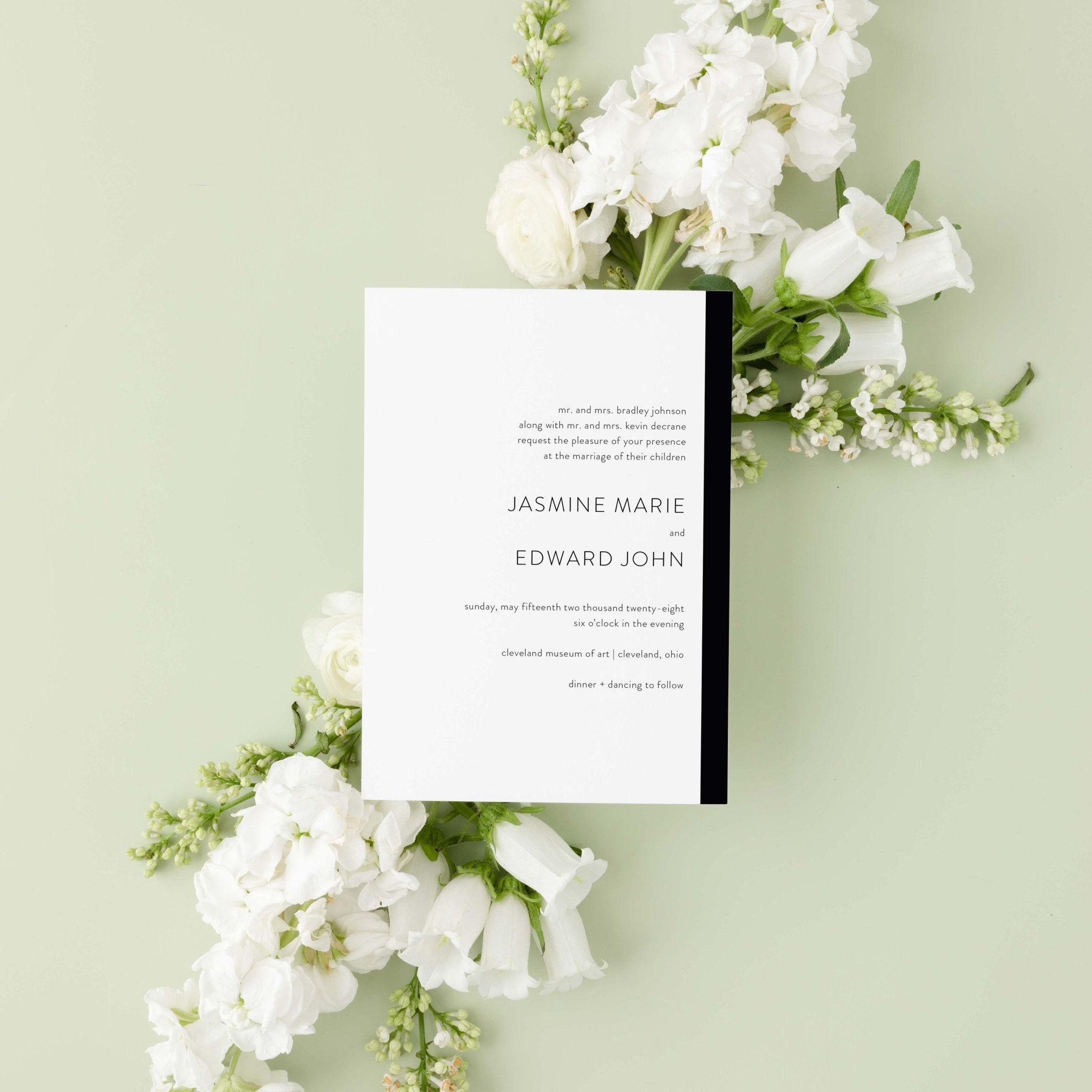 Modern Love wedding invitation by Blú Rose, white card with a black side stripe and serif names, styled on a sage background with white blooms