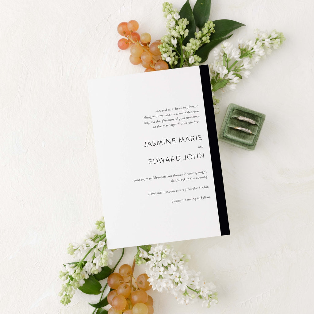 Modern Love wedding invitation by Blú Rose, white card with a black side stripe and serif names, styled with white lilacs, grapes, and wedding rings