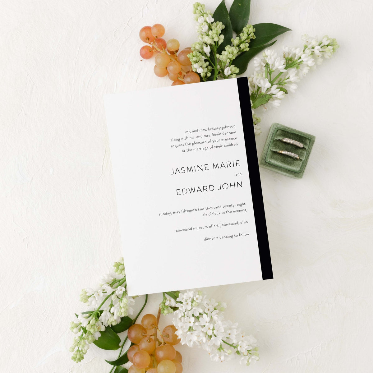 Modern Love wedding invitation by Blú Rose, white card with a black side stripe and serif names, styled with white lilacs, grapes, and wedding rings