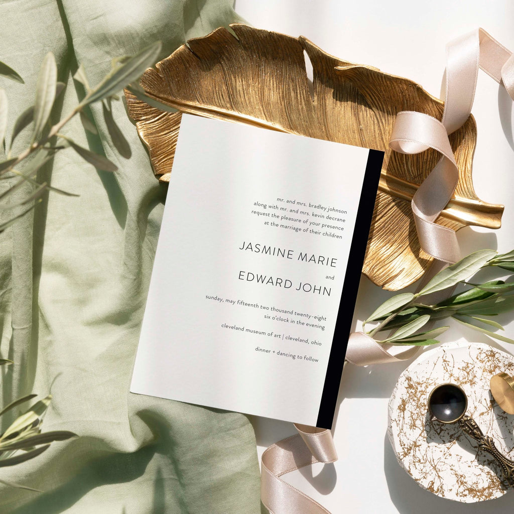 Modern Love wedding invitation by Blú Rose, white card with a black side stripe and serif names, styled on a gold leaf dish with sage linen and ribbon