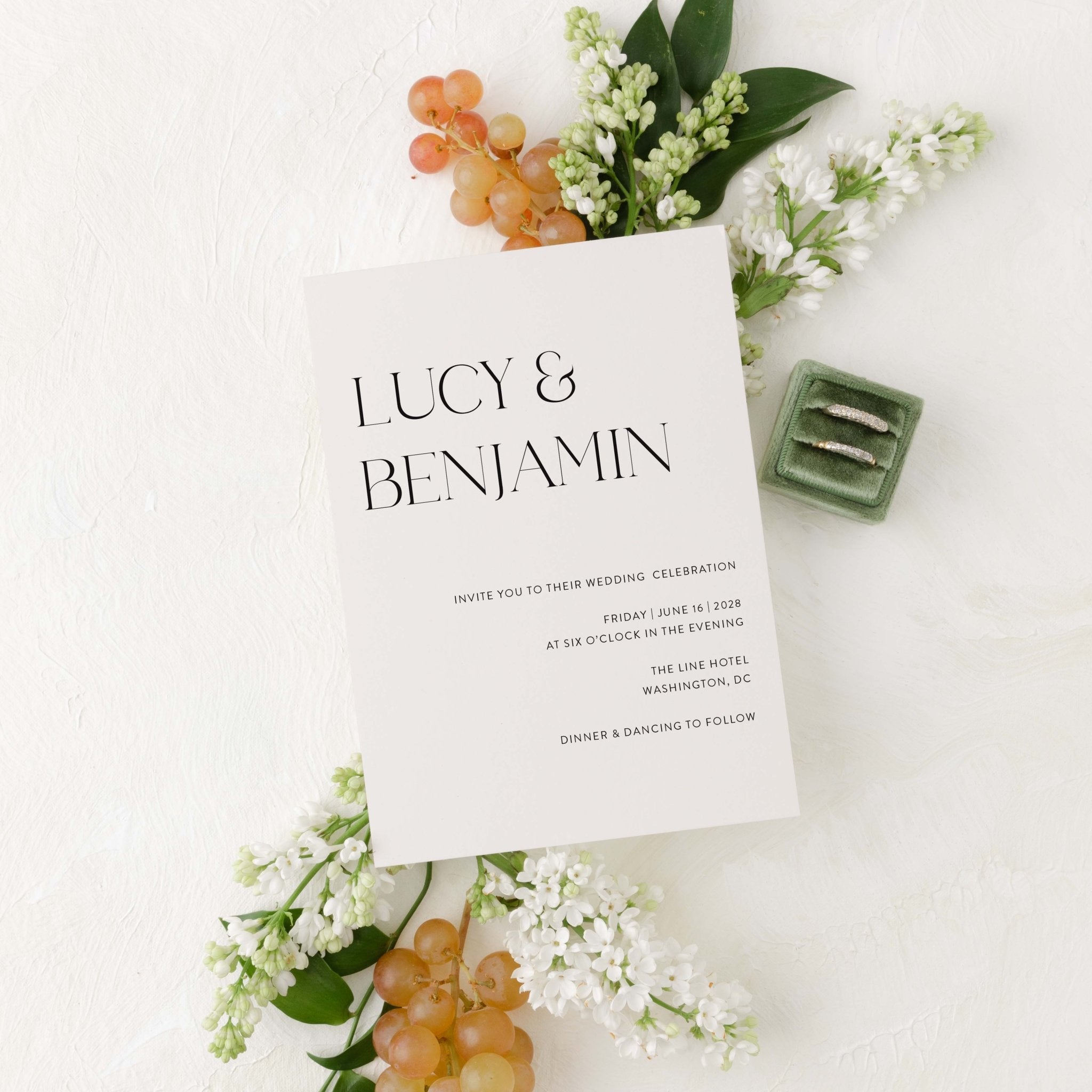 Modern Lucy wedding invitation by Blú Rose, cream card with large serif names, styled with white lilacs, grapes, and wedding rings