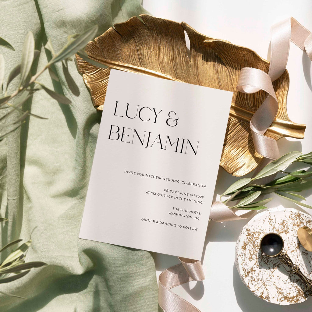 Modern Lucy wedding invitation by Blú Rose, cream card with large serif names, styled on a gold leaf dish with sage linen and ribbon