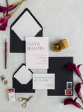 Modern Lucy wedding invitation suite by Blú Rose, minimalist invitation and reply card with large serif names, styled with black envelopes, deep red calla lilies, and a ring box