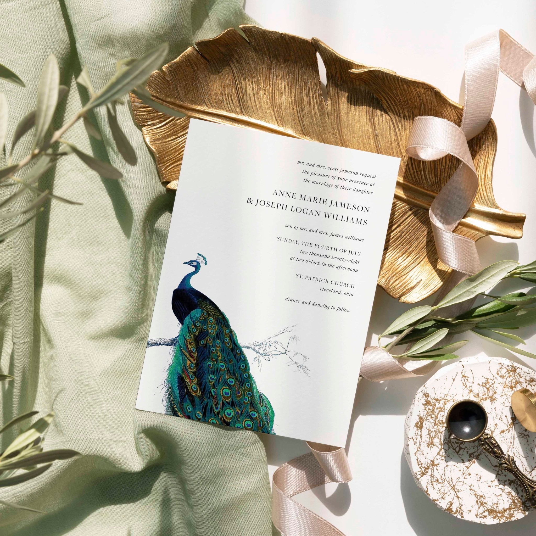 Modern Peacock wedding invitation by Blú Rose, white card with a vintage peacock illustration, styled on a gold leaf dish with sage linen and ribbon