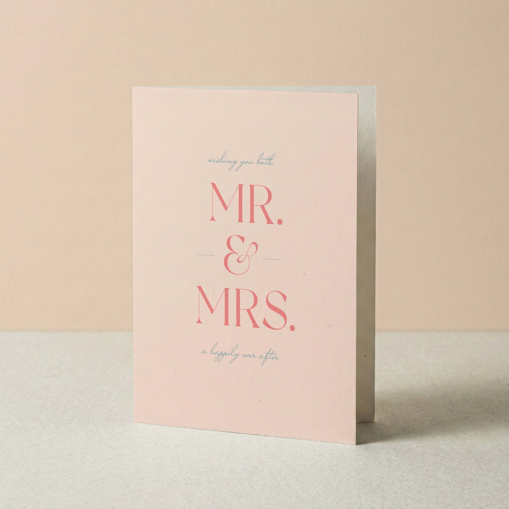 A pink greeting card standing upright on a light surface against a soft beige background. The card reads 'wishing you both MR. & MRS. a happily ever after'