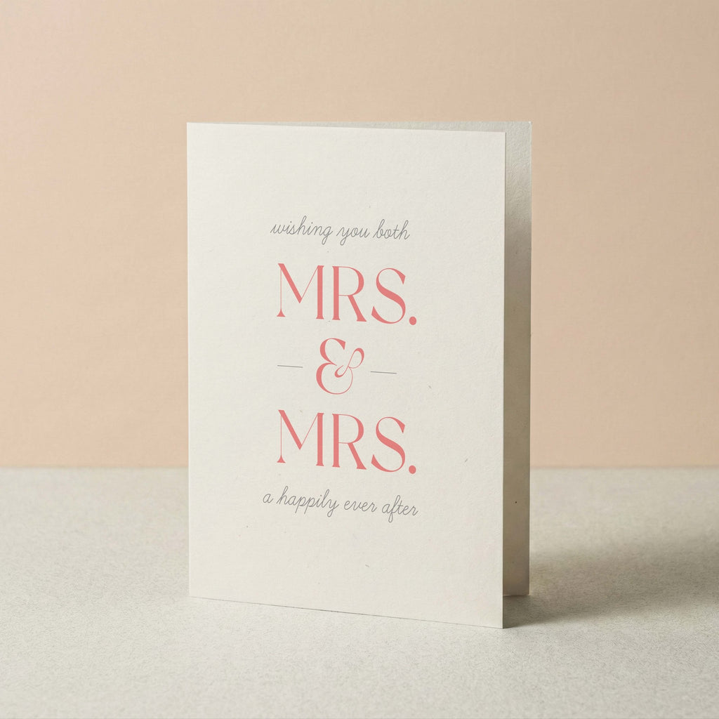 A white greeting card standing upright on a light surface against a soft peach background. The card reads 'wishing you both MRS. & MRS. a happily ever after'.