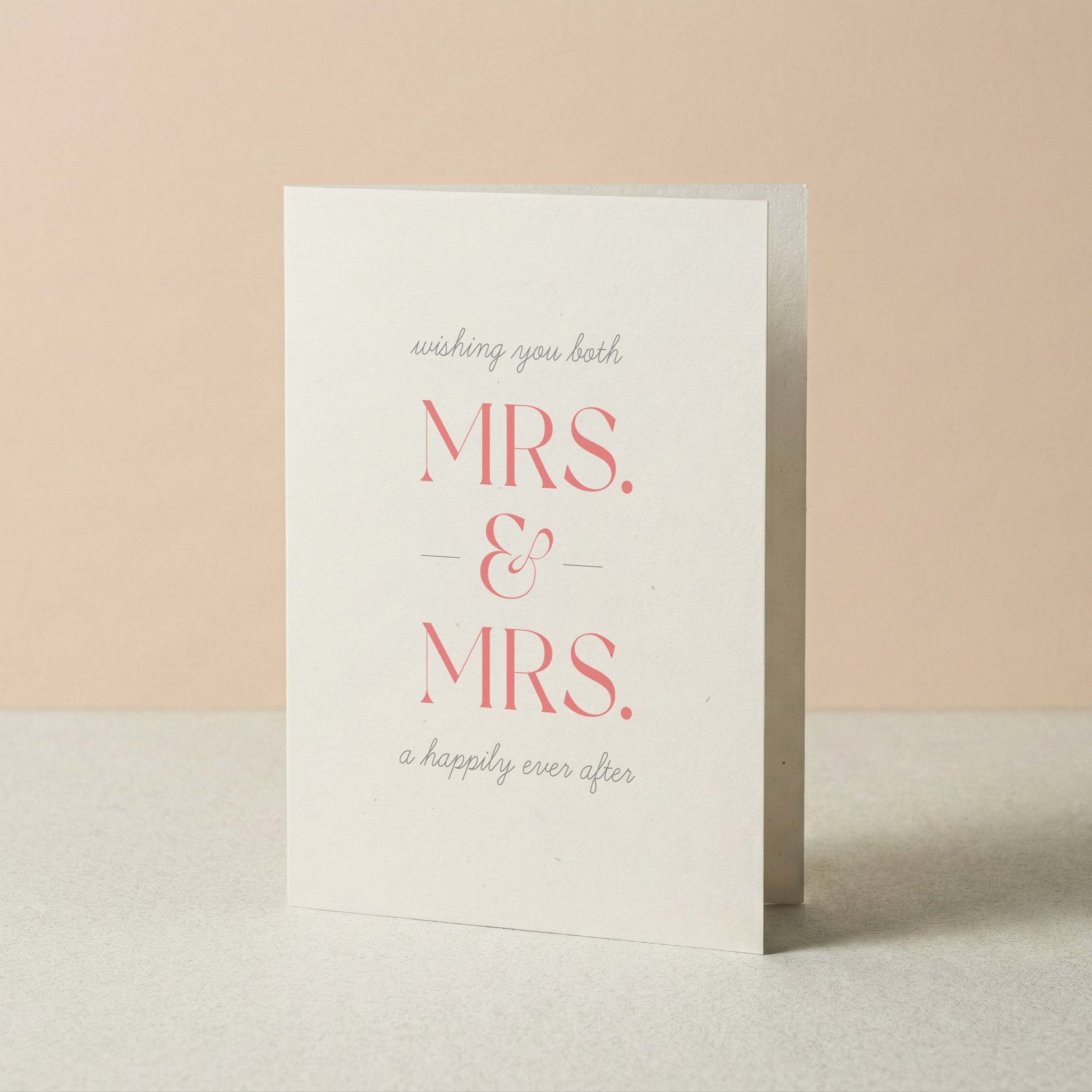 A white greeting card standing upright on a light surface against a soft peach background. The card reads 'wishing you both MRS. & MRS. a happily ever after'.
