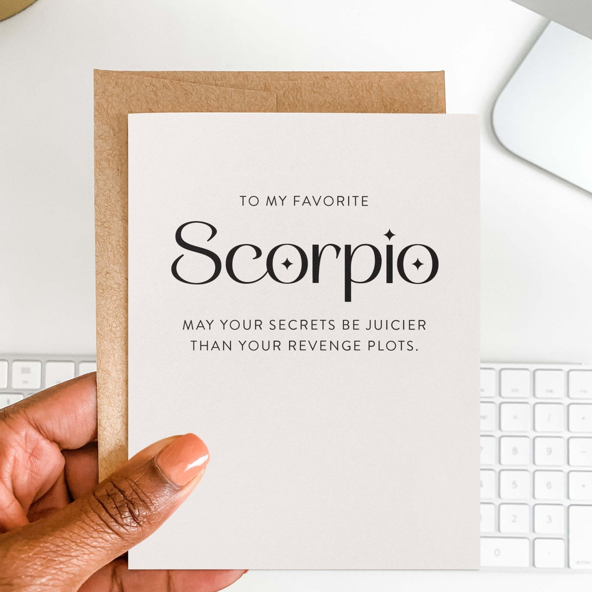 My Favorite Scorpio greeting card by Blú Rose held in hand over a kraft envelope, with the 'may your secrets be juicier than your revenge plots' design and a keyboard behind
