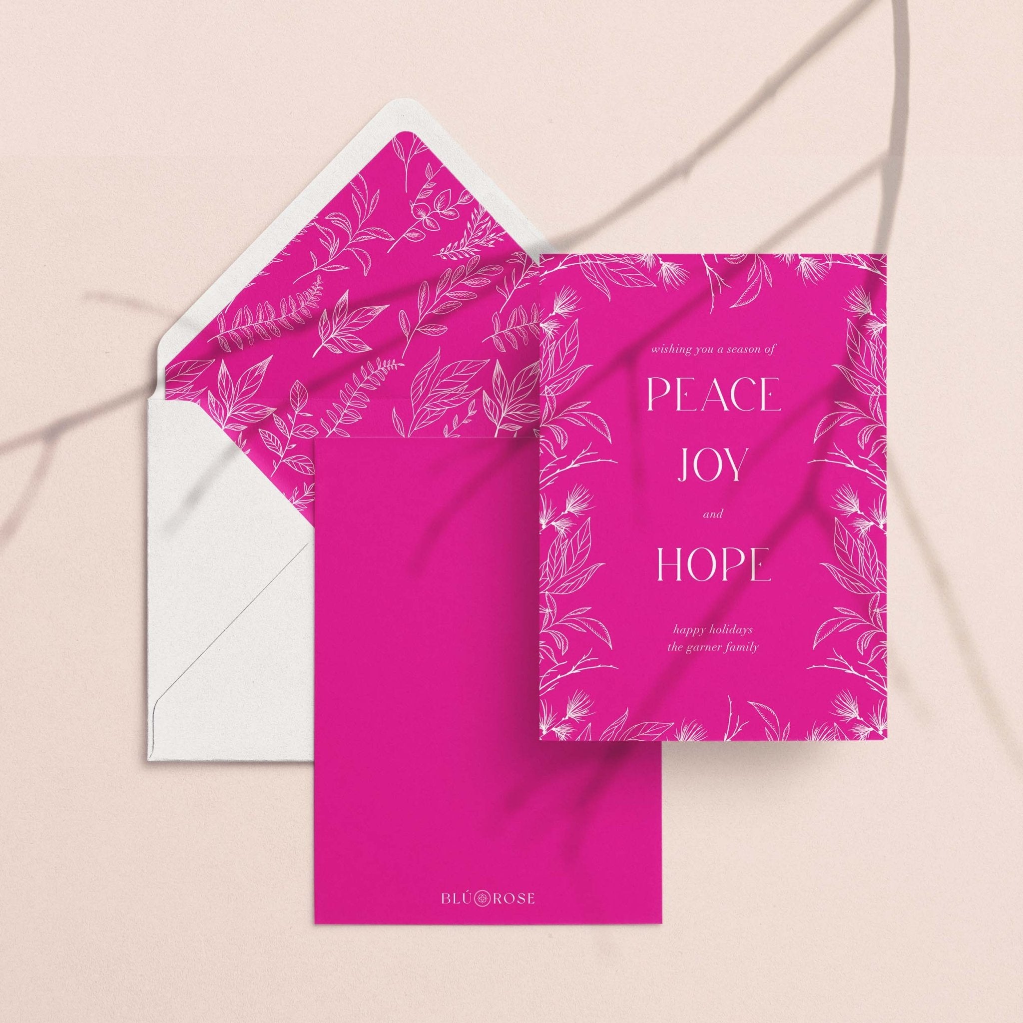Peace Joy Hope card with its magenta botanical envelope liner styled on a blush backdrop