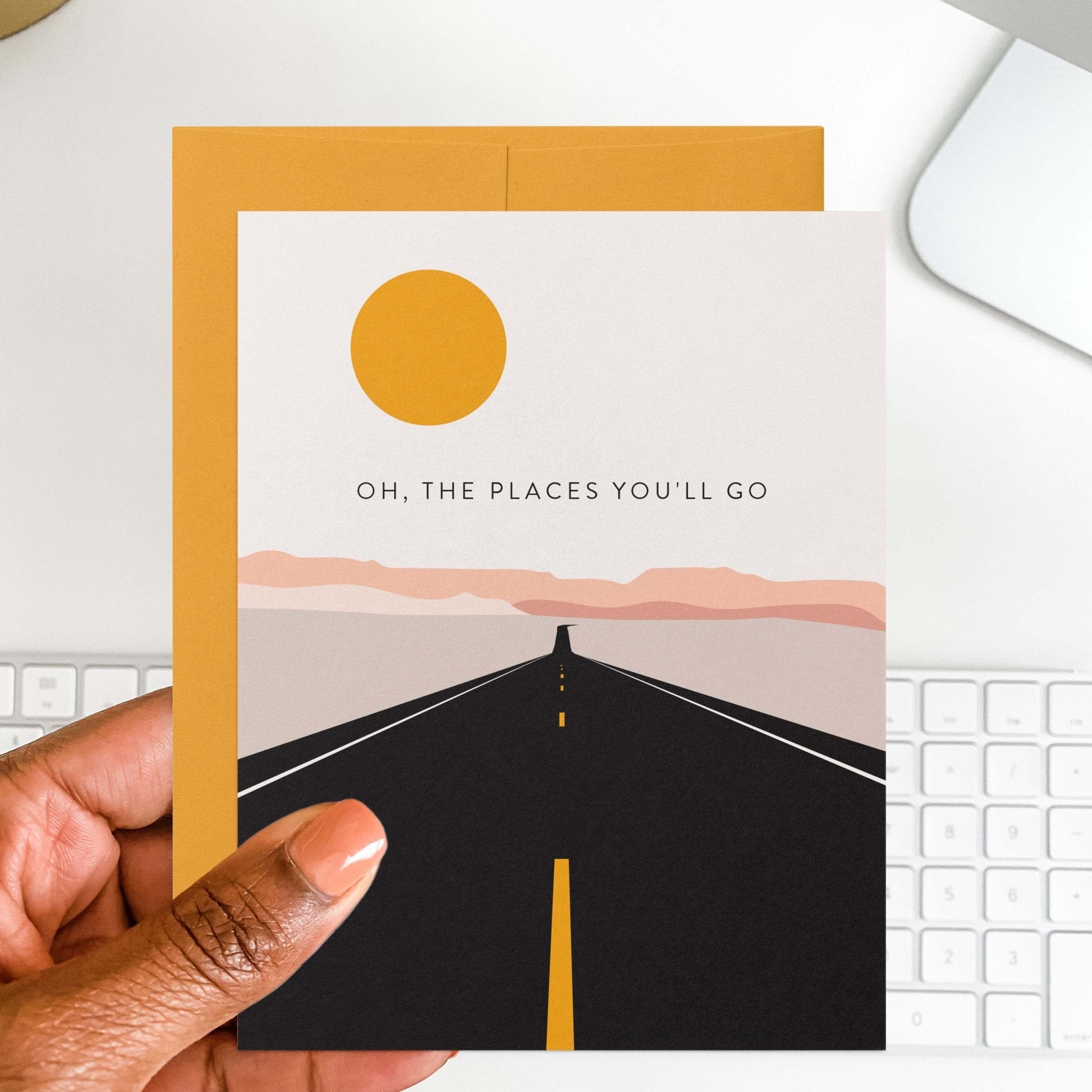 Road Trip graduation card by Blú Rose held in hand over a white desk with keyboard and marigold envelope