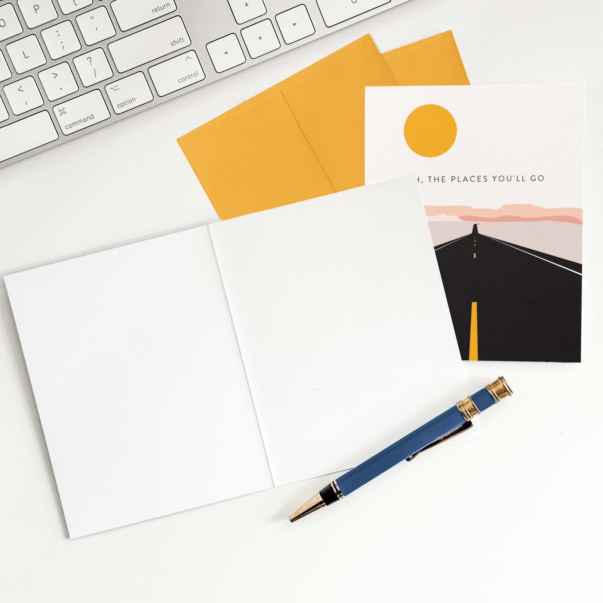 Road Trip graduation card by Blú Rose shown open with blank interior, marigold envelope and pen on a white desk