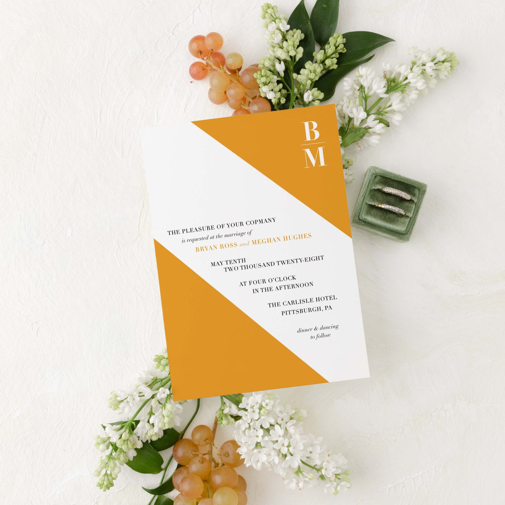 Slanted wedding invitation by Blú Rose, white card with a diagonal marigold color block and 'BM' monogram, styled with white lilacs, grapes, and wedding rings