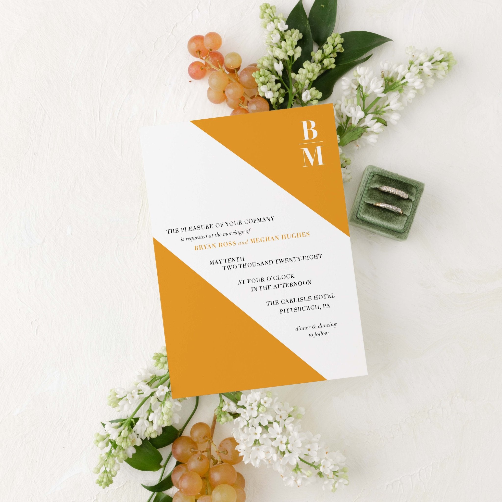 Slanted wedding invitation by Blú Rose, white card with a diagonal marigold color block and 'BM' monogram, styled with white lilacs, grapes, and wedding rings