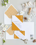 Slanted wedding invitation suite by Blú Rose, invitation and reply card with a diagonal marigold color block and monogram, styled with marigold geometric-lined envelopes, ribbon, and a wax seal