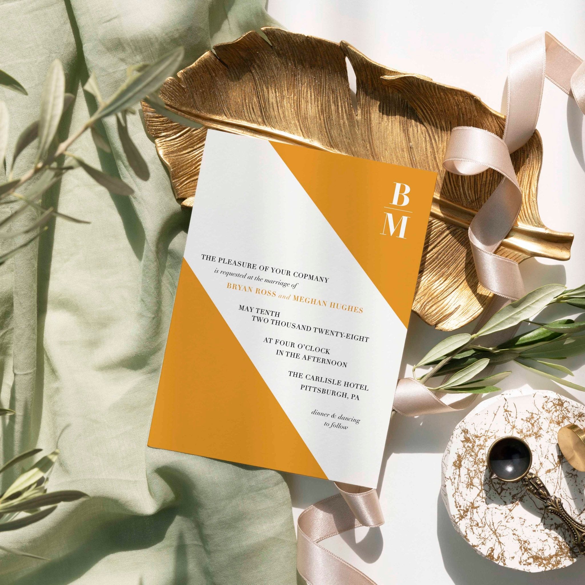 Slanted wedding invitation by Blú Rose, white card with a diagonal marigold color block and monogram, styled on a gold leaf dish with sage linen and ribbon