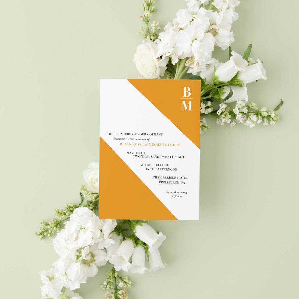 Slanted wedding invitation by Blú Rose, white card with a diagonal marigold color block and monogram, styled on a sage background with white blooms