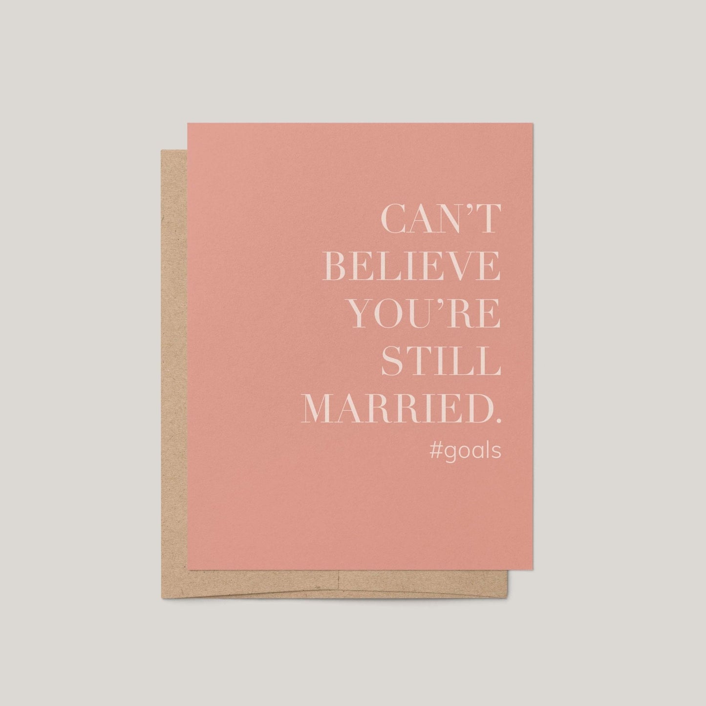 Card with text 'Can't believe you're still married. #goals' on a pink background with a brown envelope.