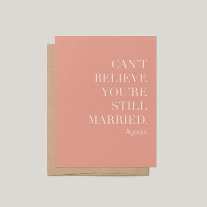 Card with text 'Can't believe you're still married. #goals' on a pink background with a brown envelope.