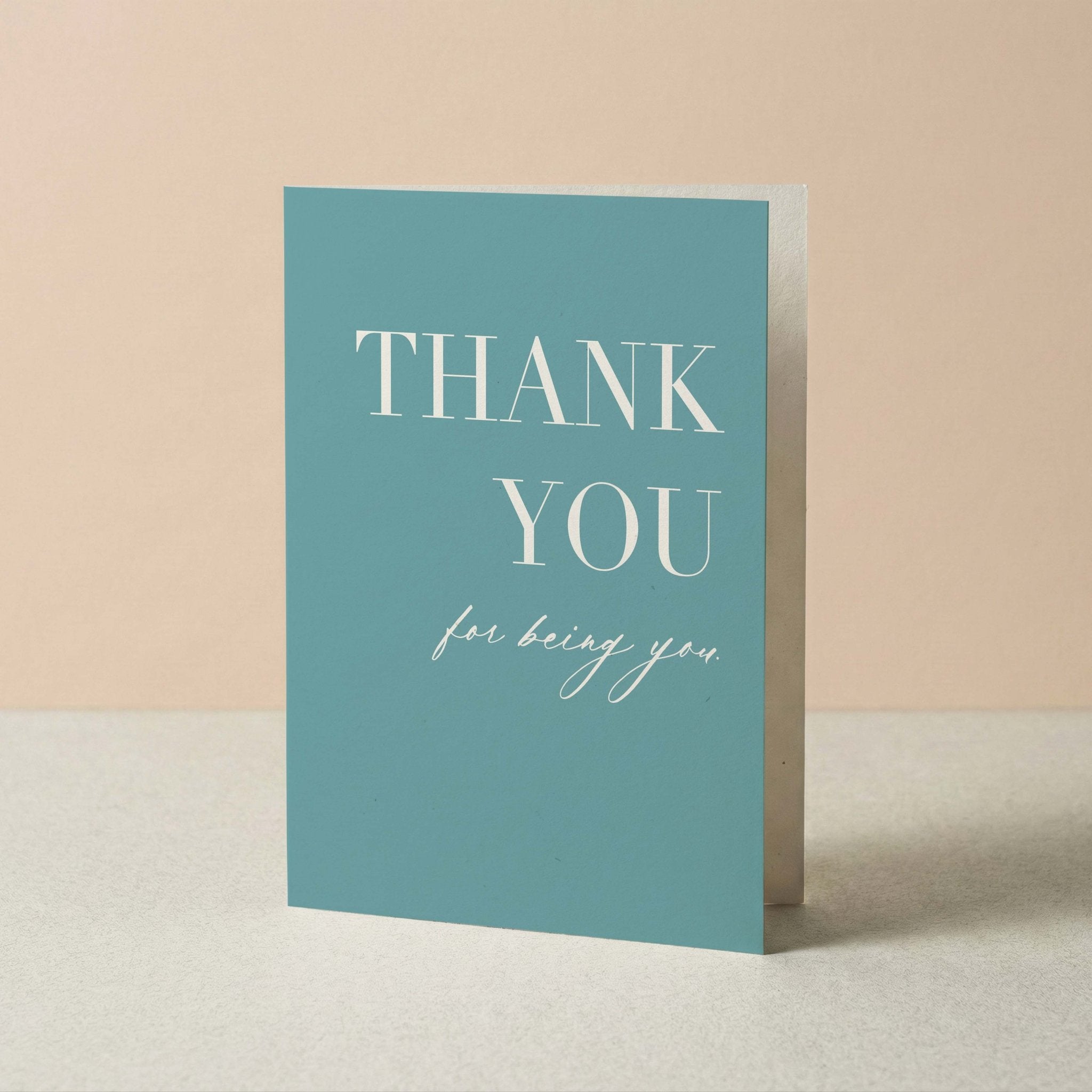A vertical teal-blue thank you card with white text reading 'THANK YOU for being you,' standing upright on a light surface against a soft beige background.