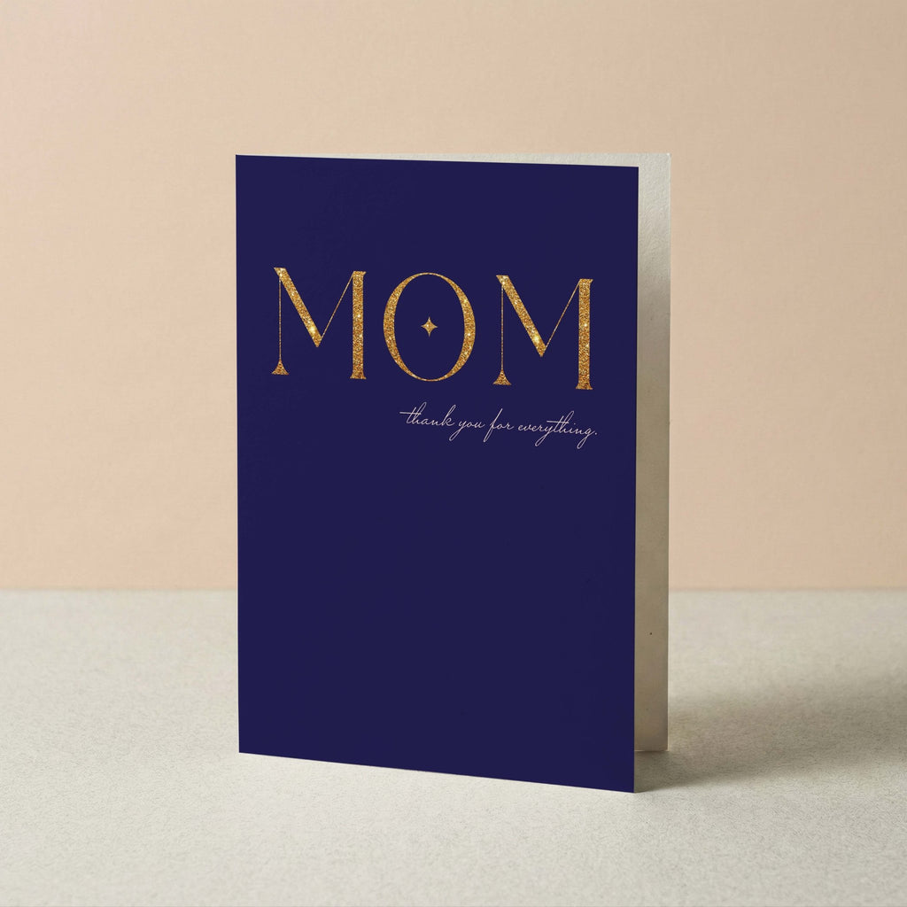 A navy blue greeting card with 'MOM' in large gold letters and the phrase 'thank you for everything' in small cursive script, standing upright on a light surface against a warm beige background.