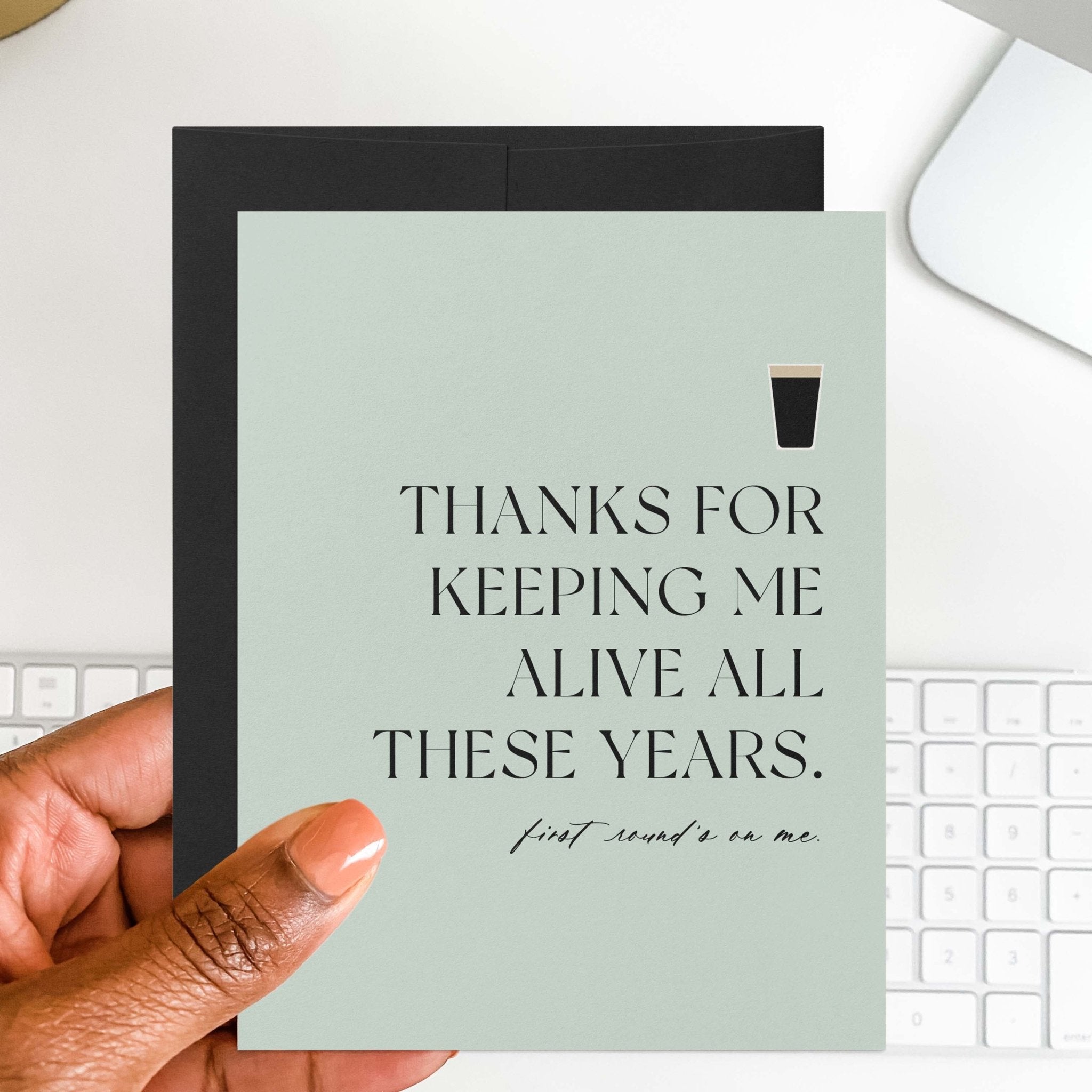Thanks for Keeping Me Alive funny Father's Day card by Blú Rose held in hand over a white desk with keyboard and black envelope