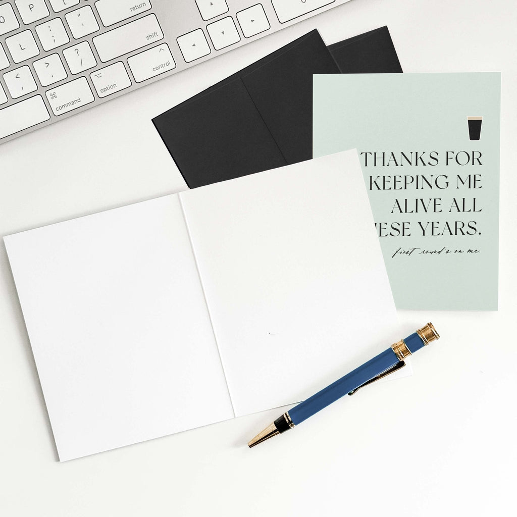 Thanks for Keeping Me Alive card by Blú Rose shown open with blank interior, black envelope and pen on a white desk