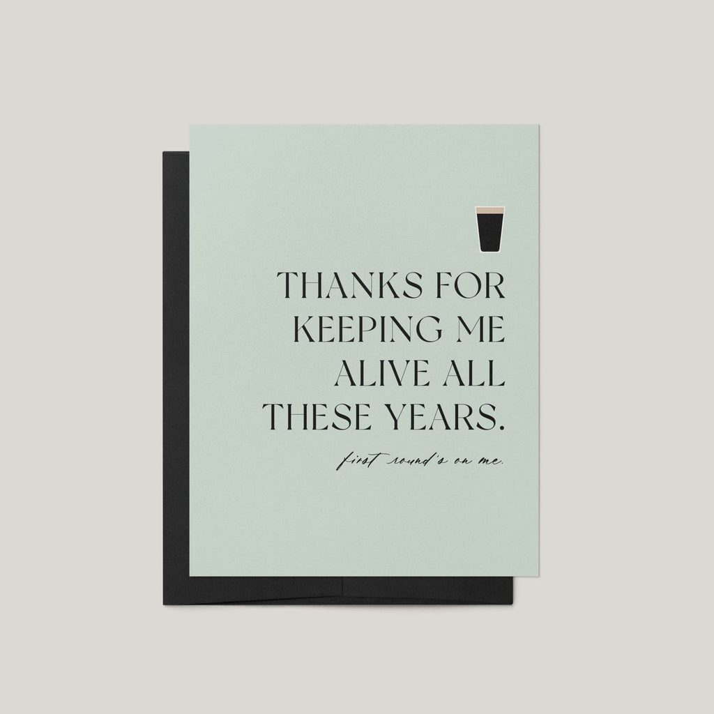 Thanks for Keeping Me Alive funny Father's Day greeting card by Blú Rose, sage card with beer pint illustration and first round's on me text, shown with black envelope