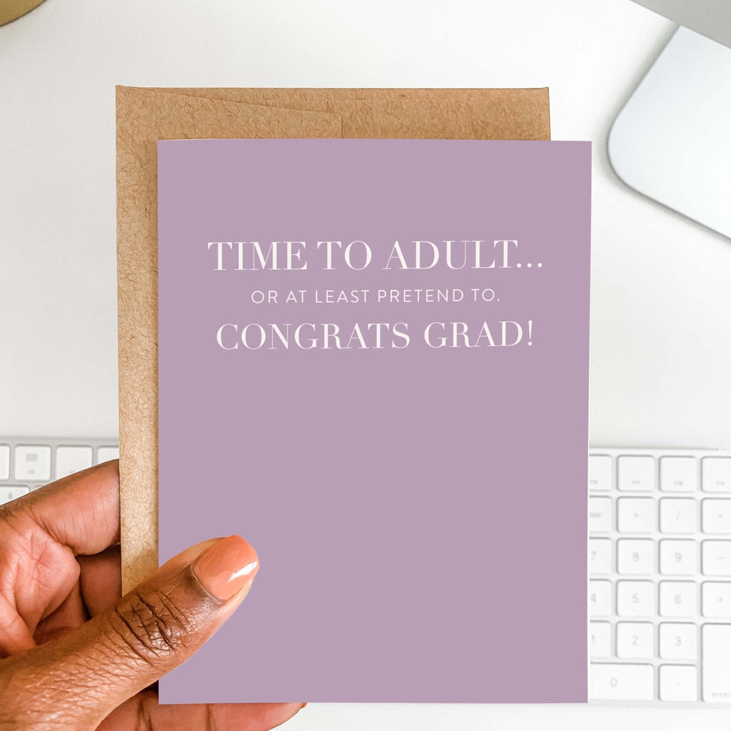 Time to Adult funny graduation card by Blú Rose held in hand over a white desk with keyboard and kraft envelope