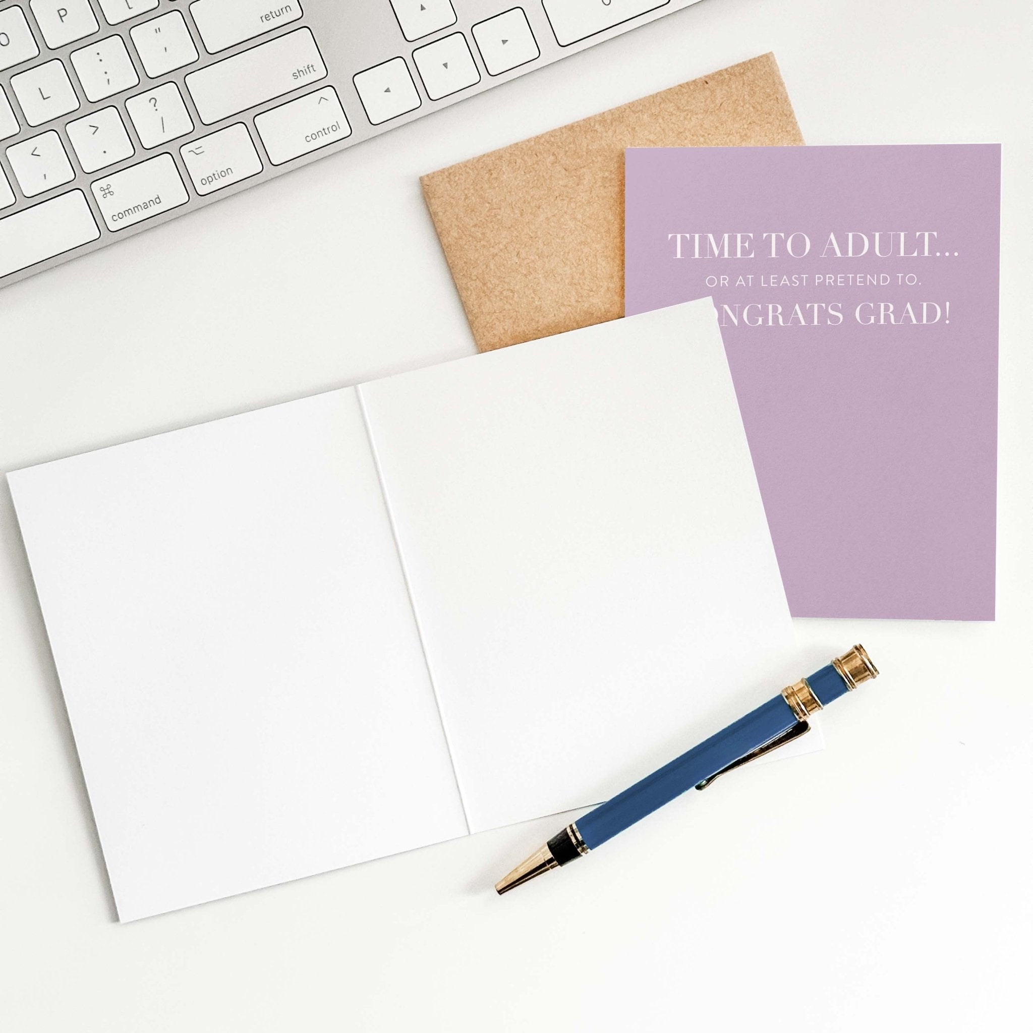 Time to Adult graduation card by Blú Rose shown open with blank interior, kraft envelope and pen on a white desk