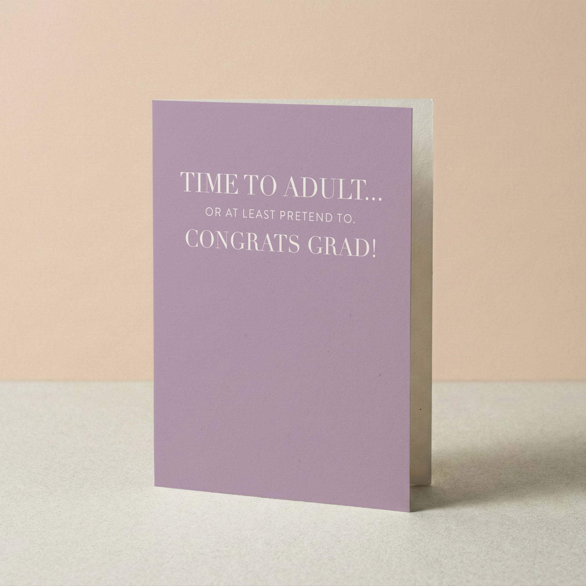 Time to Adult graduation card by Blú Rose standing upright, lavender card with congrats grad message on a neutral background