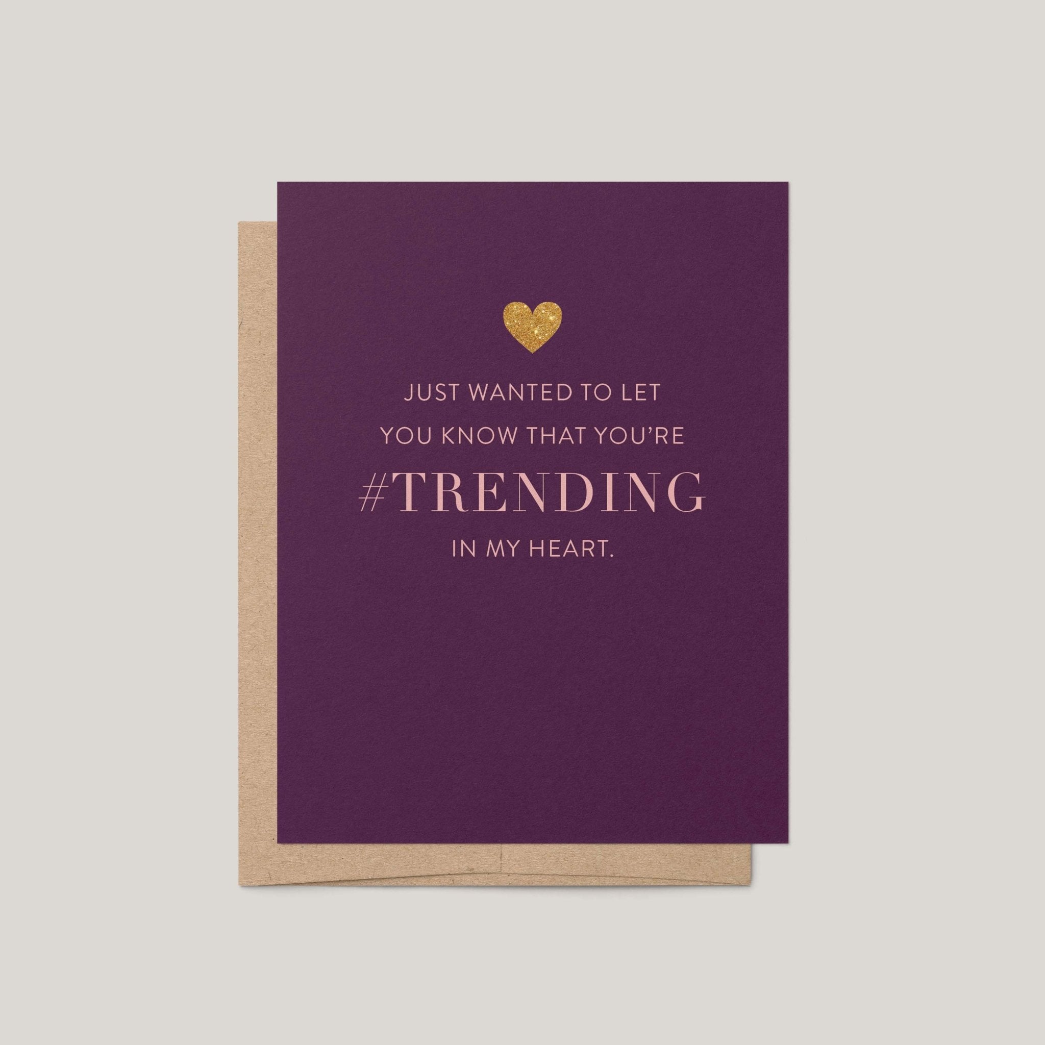 A purple greeting card with a gold heart and the text 'JUST WANTED TO LET YOU KNOW THAT YOU'RE #TRENDING IN MY HEART.' shown with a kraft envelope on a light gray background.