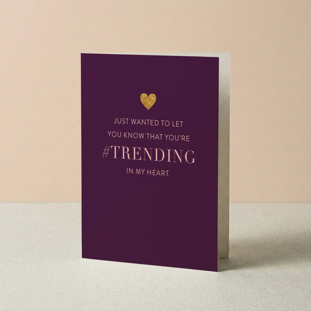 A purple greeting card with gold foil text reading 'JUST WANTED TO LET YOU KNOW THAT YOU'RE #TRENDING IN MY HEART.' and a small gold heart, standing upright against a soft beige background.