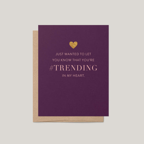 A purple greeting card with a gold heart and the text 'JUST WANTED TO LET YOU KNOW THAT YOU'RE #TRENDING IN MY HEART.' shown with a kraft envelope on a light gray background.