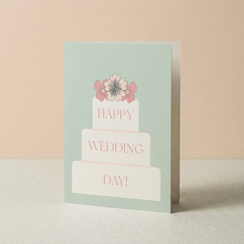 A greeting card standing upright on a light surface against a soft beige background. The card has a pale mint green border and a white tiered cake illustration with pink flowers and the words 'HAPPY WEDDING DAY!' in pink.